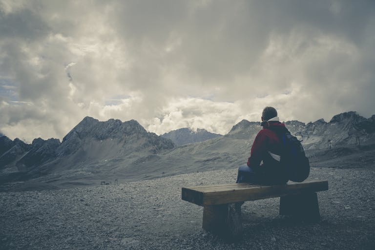 A solitary traveler sits on a bench, gazing at a mountain range under a cloudy sky.